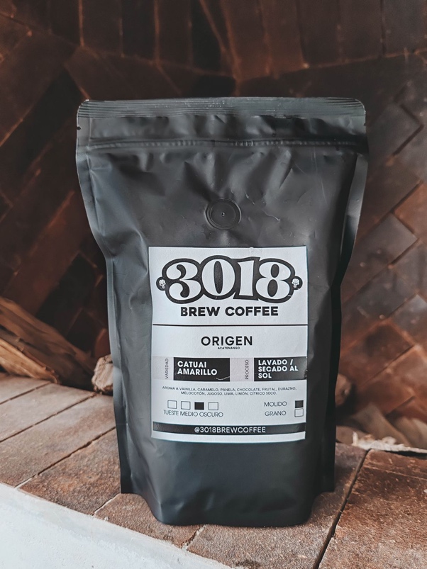 Bolsa negra de café ORIGEN de pie sobre ladrillo frente a una pared de madera; la etiqueta indica Acatenango, variedad Catuai Amarillo y proceso lavado.