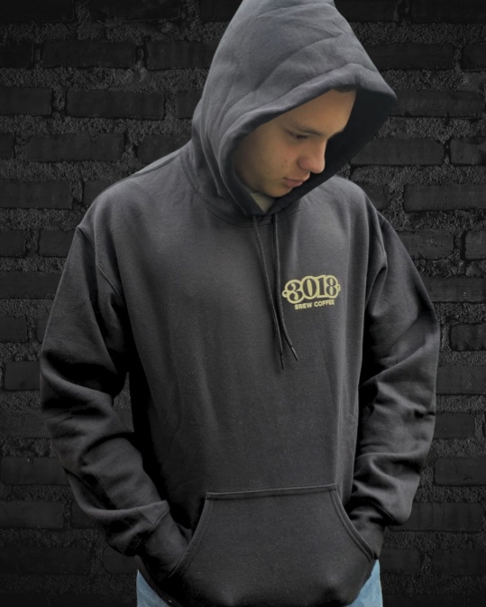 Hoodie negro puesto con la capucha arriba y el logo 3018 Brew Coffee bordado en crema sobre el pecho, frente a una pared de ladrillo negro.
